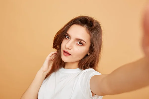 Brunette meisje met lang haar gekleed in witte t-shirt maakt een selfie op de beige achtergrond in de studio — Stockfoto