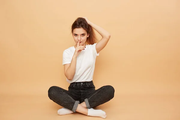 Leuk meisje gekleed in een wit t-shirt, jeans en witte sokken zit op de vloer met haar vinger op haar lippen op de beige achtergrond in de studio — Stockfoto