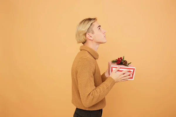 Young blond guy dressed in mustard color sweater holds a Christmas gift in his hands on the beige background in the studio