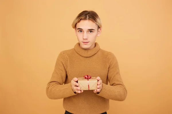 겨자 색 스웨터를 입은 젊은 금발 남자는 크리스마스 선물 스튜디오에서 베이지색 배경에 그의 손에 보유 하 — 스톡 사진