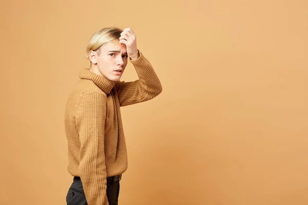 Giovane ragazzo biondo vestito in maglione color senape e pantaloni neri è in posa con la mano sulla fronte in studio sullo sfondo beige — Foto Stock