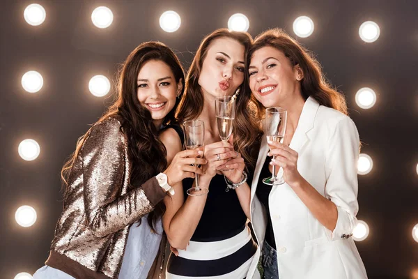 Üç güzel kız şık rahat kıyafetler içinde bardak şampanya el ışıklar arka plan duvar üzerinde gülümseyen tutun. Parti zamanı