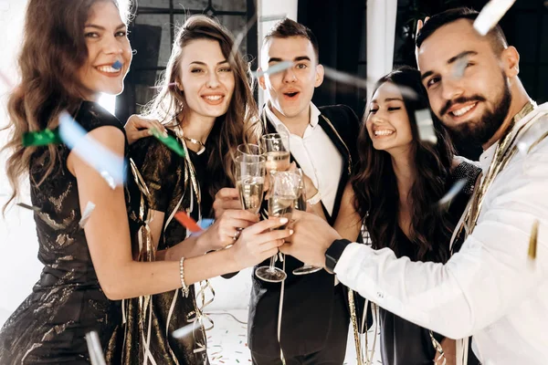 Hermosas chicas jóvenes y chicos vestidos con ropa elegante y elegante sonríen juntos y tintinean copas con champán. Tiempo de fiesta — Foto de Stock