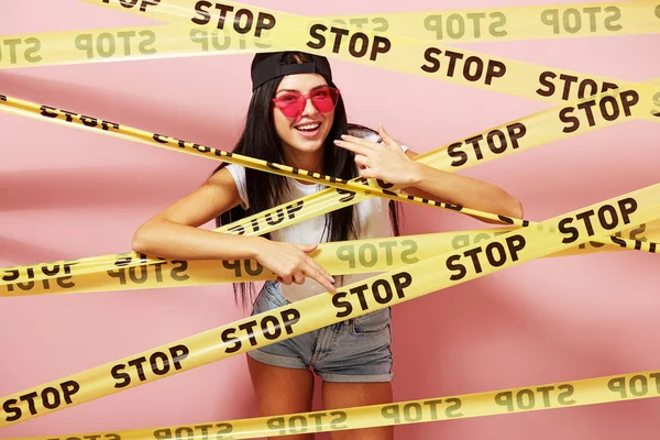 Χαμογελώντας μελαχρινός κορίτσι στο ροζ γυαλιά ηλίου και καπάκι κοιτάζοντας έξω από πίσω από κίτρινο ταινίες scotch με επιγραφές της λέξης "stop" στο φόντο των ροζ τοίχο — Φωτογραφία Αρχείου