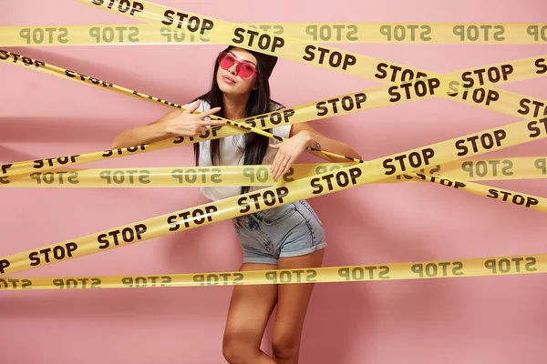 Dunkelhaariges Mädchen mit pinkfarbener Sonnenbrille und Mütze, bekleidet mit Jeanshosen und weißem Oberteil, das hinter gelben Schottenbändern mit der Aufschrift "Stop" auf dem Hintergrund der rosa Wand hervorschaut — Stockfoto