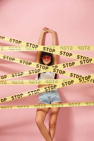 Mladá černovlasá dívka v růžové brýle a čepici oblečený v džínách šortky a bílé nejlepší stojí za žluté pásky scotch s nápisy slovem "stop" na pozadí růžové stěny — Stock fotografie