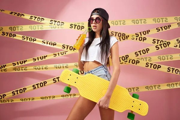 Szczupła dziewczyna w różowe okulary przeciwsłoneczne i czapka ubrany w dżinsy szorty i biały top stoi żółty deskorolka i butelka soku na tle muru różowy z taśm scotch z słowo "stop" — Zdjęcie stockowe