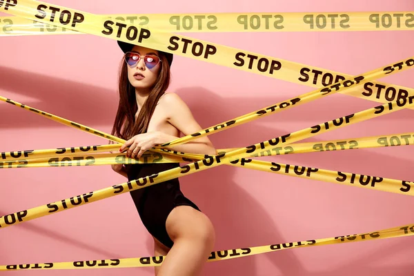 Schlankes Mädchen mit Sonnenbrille und Mütze in schwarzem Badeanzug, das hinter gelben Schottenbändern mit der Aufschrift "Stop" auf dem Hintergrund der rosafarbenen Wand hervorblickt — Stockfoto