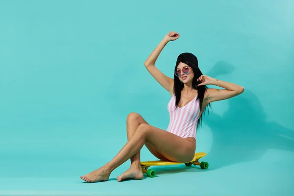 Menina de cabelos escuros esbeltos em óculos de sol e boné vestido com maiô listrado rosa e branco senta-se no skate amarelo no fundo azul no estúdio — Fotografia de Stock