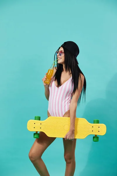 Slim dark-haired girl in sunglasses and cap dressed in pink and white striped swimsuit stands with yellow skateboard and bottle of juice in her hands on the blue background — Stock Photo, Image