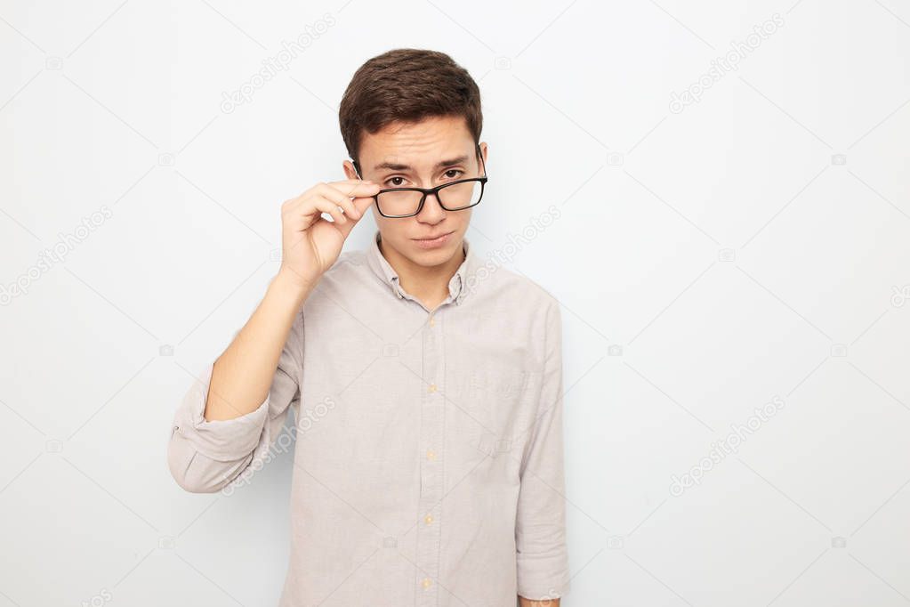 Young guy in glasses dressed in light shirt stands with his hand on his glasses on the white background in the studio