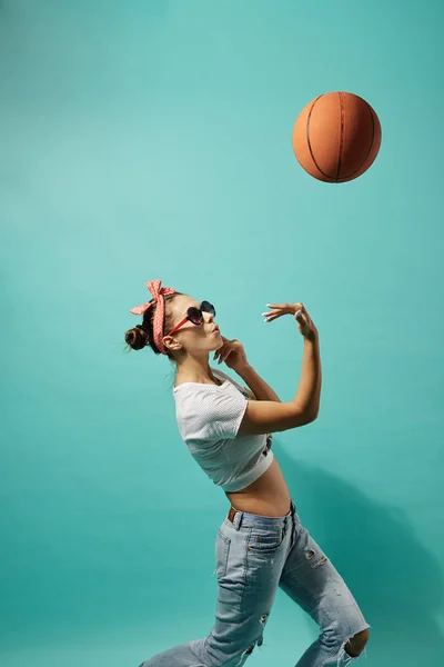 Jovencita con gafas de sol y lazo rosa en la cabeza vestida con jeans y top lanza una pelota de baloncesto sobre el fondo azul en el estudio — Foto de Stock