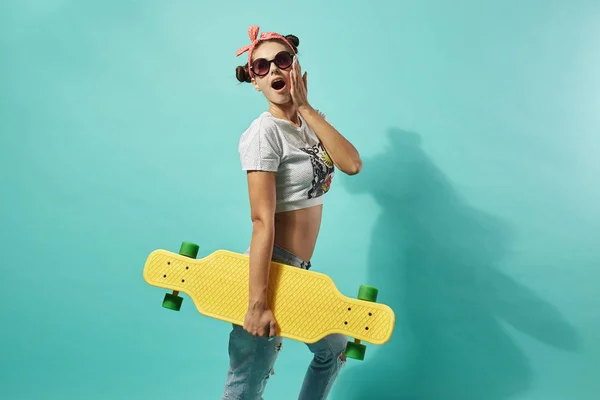 Grappige meisje in zonnebrillen en roze strik op haar hoofd gekleed in jeans en top stands met gele skateboard op de rug op de blauwe achtergrond in de studio — Stockfoto