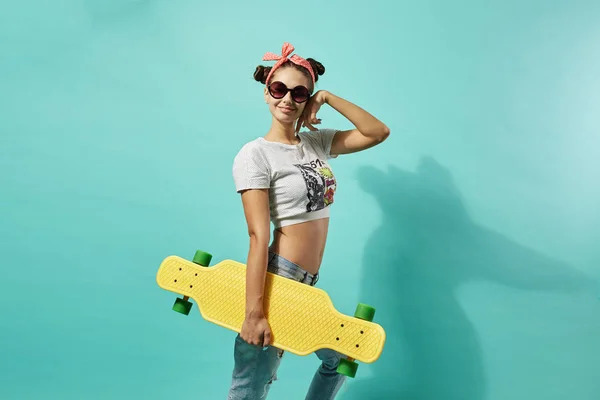 Engraçado jovem em óculos de sol e laço rosa na cabeça vestida de jeans e top stands com skate amarelo na parte de trás no fundo azul no estúdio — Fotografia de Stock