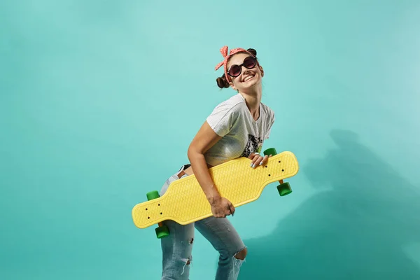 Zabawna dziewczyna w okulary przeciwsłoneczne i różowy kokardkę na głowie, ubrany w dżinsy i najlepsze Stoiska z żółtym deskorolka tyłu na niebieskim tle w studio — Zdjęcie stockowe