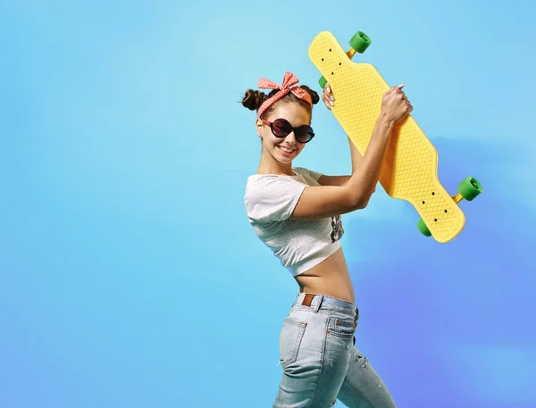 Young girl in sunglasses and pink bow on her head dressed in jeans and top stands with yellow skateboard on the back on the blue background in the studio — Stock Photo, Image