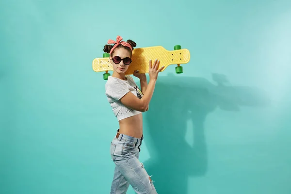 선글라스와 청바지와 스튜디오에서 파란색 배경에 뒤쪽에 노란 스케이트 보드와 최고의 스탠드 입고 그녀의 머리에 분홍색 활에 재미 있는 어린 소녀 — 스톡 사진