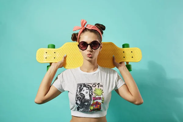 Lustiges junges Mädchen mit Sonnenbrille und rosa Schleife auf dem Kopf, bekleidet mit Jeans und Top-Ständer mit gelbem Skateboard auf dem Rücken auf blauem Hintergrund im Studio — Stockfoto