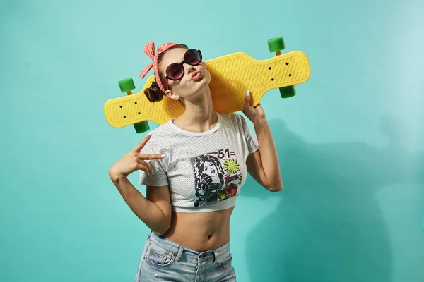 Joyeux jeune fille en lunettes de soleil et arc rose sur sa tête vêtue de jeans et haut se tient avec skateboard jaune sur le fond bleu dans le studio — Photo