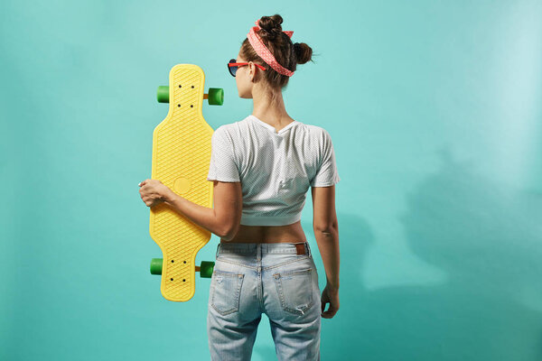 Back view of a girl dressed in jeans and top stands with yellow skateboard on the blue background in the studio