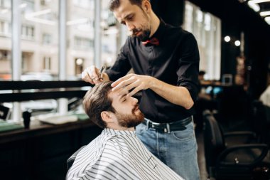 Kuaför kırmızı papyon ile siyah bir gömlek giymiş bıyıklı bir berber dükkanı genç adam için şık bir saç modeli yapar