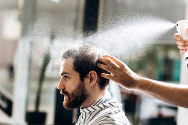 Bir siyah gömlek kırmızı papyon ve düz kapak giymiş bıyıklı Kuaför saç şekillendirme bir berber dükkanı koltukta oturan adam için ne yapıyor