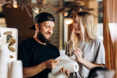 İki genç gülümseyen insanlar, bir sarışın kız ve gündelik kıyafeti, beard.dressed adamla yan yana durmak ve rahat bir kafede bakıyoruz.