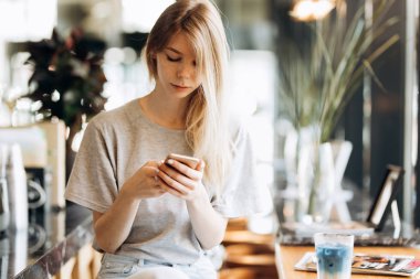 Casual tarzda giyinmiş bir sevimli ince sarışın kız, kahve içer ve telefonunu bir kafede bakar.