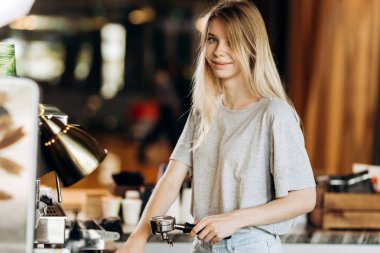 Gündelik kıyafeti giymiş bir şirin sarışın youg kız, kahve makinesi ve gülümseme rahat bir kafede sonraki standları.
