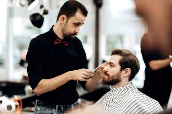 Стильный усатый парикмахер, одетый в черную рубашку с красными ножницами бабочки волосы молодого человека в парикмахерской — стоковое фото