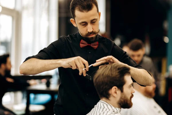 Харизматичный парикмахер с усами, одетый в черную рубашку с красными ножницами бабочки волосы молодого человека в парикмахерской — стоковое фото