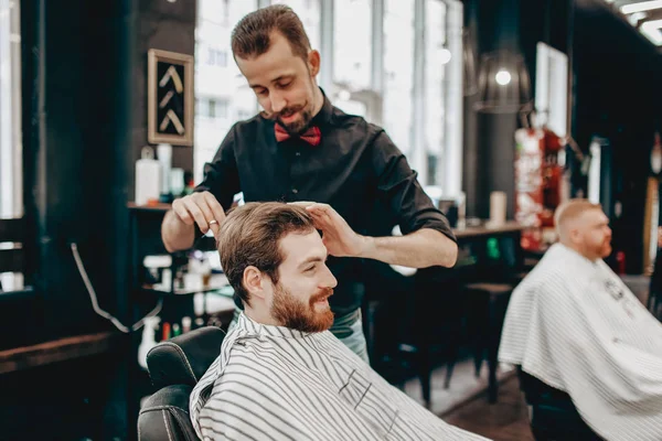 Мусташный парикмахер в черной рубашке с красной бабочкой делает стильную прическу молодому человеку в парикмахерской — стоковое фото