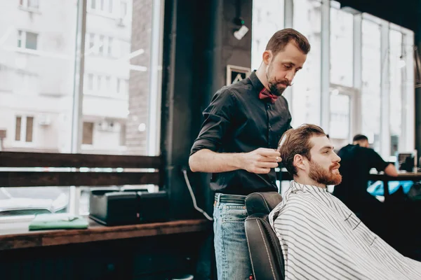 Мусташный парикмахер в черной рубашке с красной бабочкой делает стильную прическу молодому человеку в парикмахерской — стоковое фото