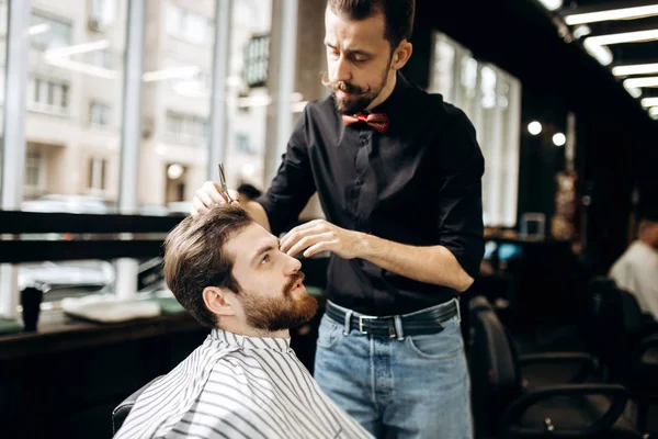 Парикмахер с усами, одетый в черную рубашку с красной бабочкой, делает стильную прическу молодому человеку в парикмахерской — стоковое фото