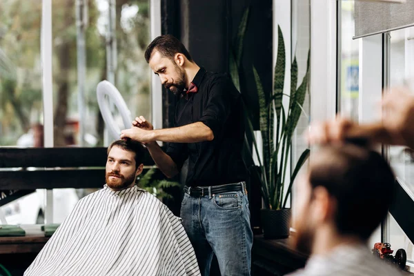 Barber dressed in a black shirt with a red bow tie scissors the mans hair in a barbershop