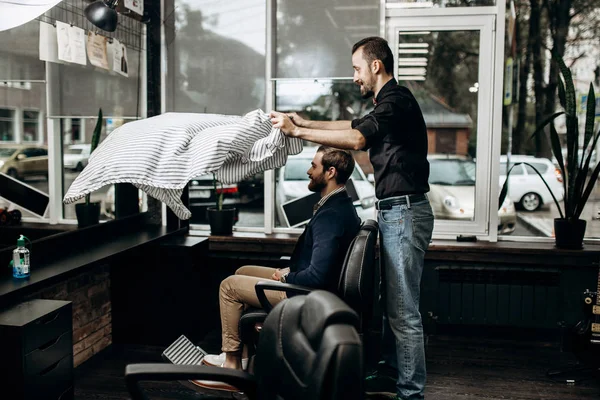 Barbeiro mustachioed vestido com uma camisa preta coloca uma capa sobre o homem elegante sentado na poltrona em uma barbearia — Fotografia de Stock