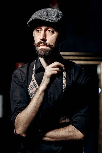 Barbeiro de moda com bigode e barba em um boné plano e camisa preta com gravata borboleta desatada fica no quarto escuro em uma barbearia — Fotografia de Stock