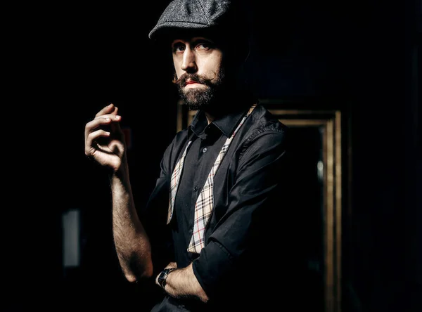 Barbier élégant avec moustache et barbe vêtu d'une chemise noire avec nœud papillon délié et casquette plate se tient dans la pièce sombre dans un salon de coiffure — Photo