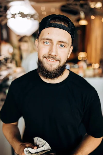 Ein gutaussehender, jugendlich stilvoller Mann mit Bart, lässig gekleidet, steht in einem gemütlichen Café und blickt in die Kamera. — Stockfoto