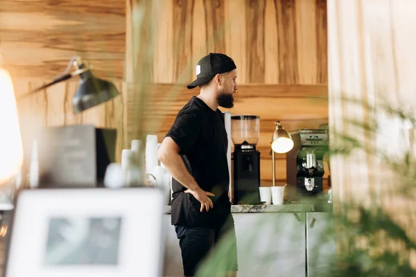 Молодий стильний чоловік з бородою, одягнений у повсякденний одяг, стоїть у затишній кав'ярні . — стокове фото