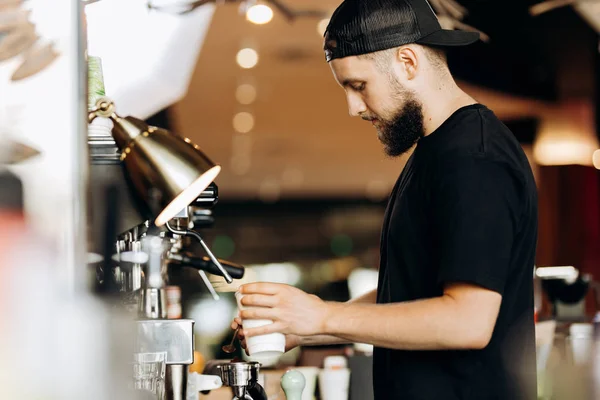 Стильний молодий чоловік з бородою, одягнений в повсякденний одяг, готує каву в кавовій машині в сучасному кафе . — стокове фото