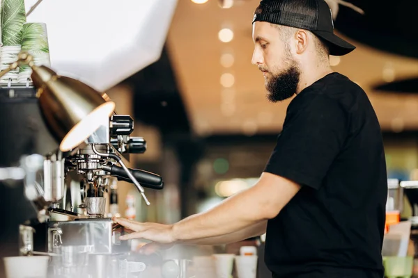 Стильний молодий чоловік з бородою, носить повсякденний одяг, готує каву в кавовій машині в затишній кав'ярні . — стокове фото