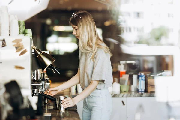 A young pretty thin blonde with long hair,dressed in casual outfit,is cooking coffee in a modern coffee shop. Process of making coffee is shown.