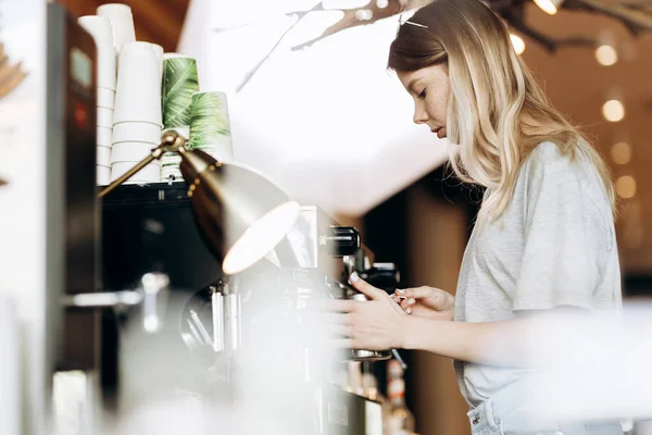 Гарна струнка блондинка з довгим волоссям, одягнена в повсякденне вбрання, готує каву в сучасному кафе. Показано процес приготування кави . — стокове фото