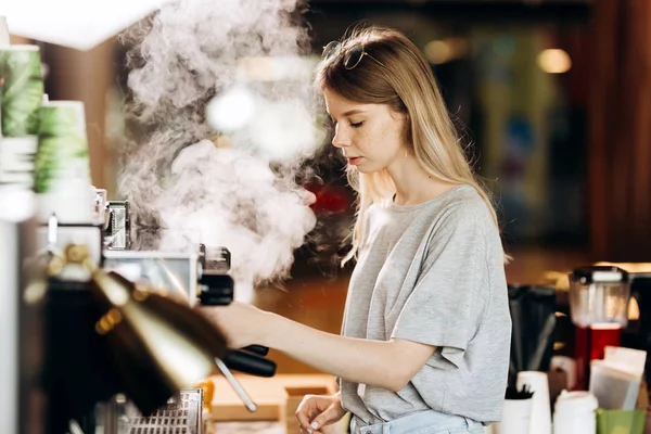 Молодий блондинка стрункий пояс, одягнений у повсякденне вбрання, очищає кавоварку парою в сучасному кафе . — стокове фото
