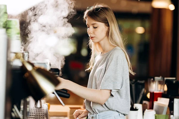 Eine jugendliche blonde schlanke Frau in lässigem Outfit putzt in einem modernen Café die Kaffeemaschine mit Dampf.. — Stockfoto