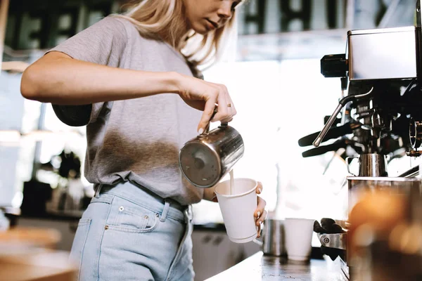 A youthful thin blonde girl,wearing casual cothes,is shown adding milk to the coffee in a cozy coffee shop.