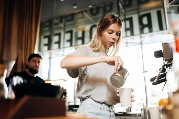 Ein jugendliches, dünnes blondes Mädchen in lässiger Kleidung wird in einem gemütlichen Café gezeigt, wie es Milch in den Kaffee gießt. — Stockfoto