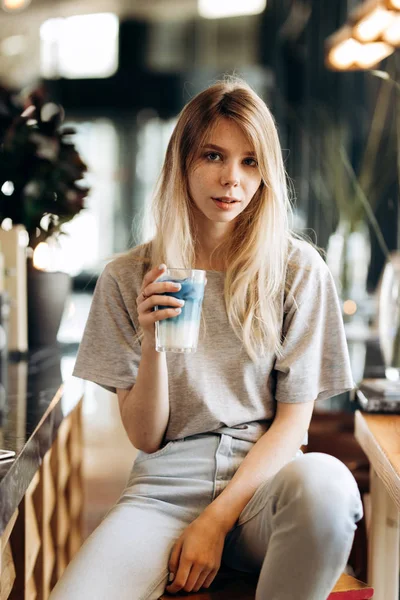 Een mooi slank meisje, het dragen van casual stijl, zit op de stoel en houdt een kop koffie in een gezellige coffee shop. — Stockfoto