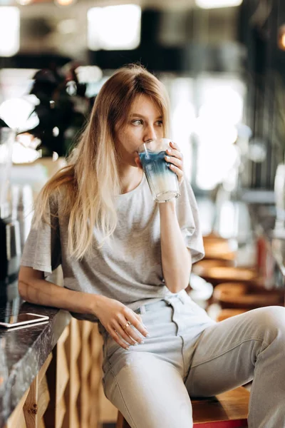 Мила струнка блондинка з довгим волоссям, одягнена в повсякденний одяг, п'є каву і дивиться на вікно . — стокове фото